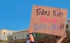 Person holding a protest sign reading ‘Trans Kids’ outside El Rio’s annual block‑party fundraiser—highlighting the El Rio Tucson Controversy and community outrage.